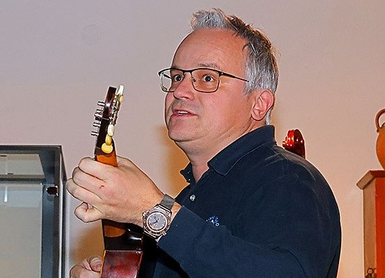 Ein Mann mit Brille und einer Uhr steht und hält eine Gitarre. Hinter ihm ist eine weiße Wand mit einer hölzernen Vitrine.