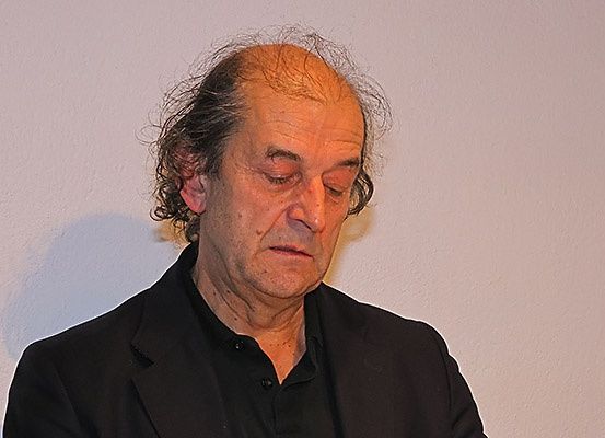 Ein älterer Mann mit grauem Haar und einem Schnurrbart sitzt mit geschlossenen Augen in einem schwach beleuchteten Raum. Er trägt ein schwarzes Hemd.