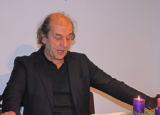 Ein älterer Mann sitzt an einem Tisch mit Kerzen darauf. Er trägt einen schwarzen Anzug und hat ein Mikrofon in der Hand. Er singt wahrscheinlich.