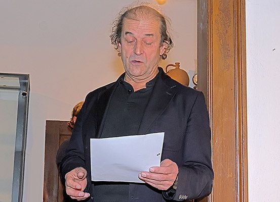 Ein älterer Mann liest mit geschlossenen Augen ein Papier, das er in einer Hand hält, in einem Raum mit Holzwänden und einer Glasscheibe.