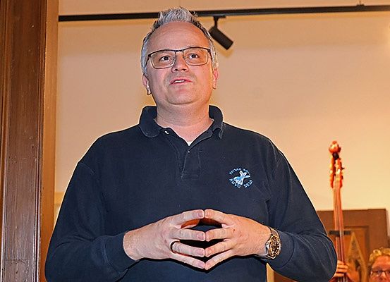 Ein Mann in einem blauen Poloshirt bildet mit seinen Händen eine Herzform und steht vor einer Holztür. Dahinter ist ein Cello und eine weitere Person zu sehen.