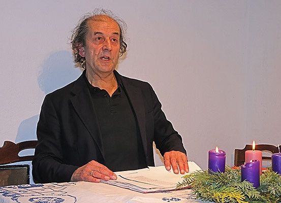 Ein älterer Mann sitzt an einem Tisch und liest ein Buch. Er ist von lila Kerzen umgeben.