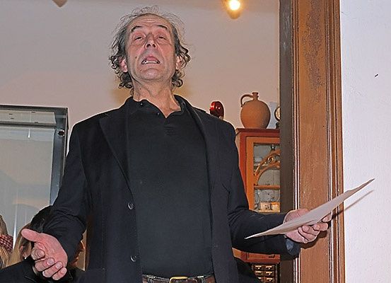Ein älterer Mann in einem schwarzen Mantel steht vor einer Wand und hält ein Stück Papier in der Hand, während ein anderer Mann hinter ihm steht.