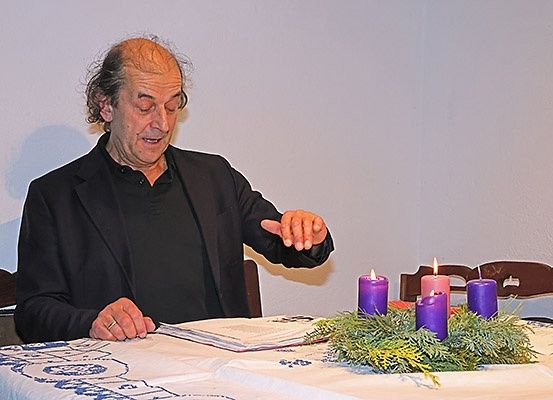 Ein älterer Mann in einem schwarzen Anzug sitzt lächelnd an einem Tisch mit Kerzen und einem offenen Buch.