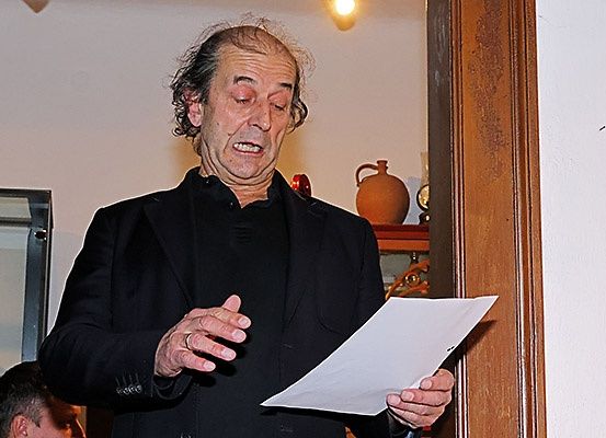 Ein Mann in einem schwarzen Anzug steht und liest ein Blatt Papier, wobei ein anderer Mann teilweise hinter ihm zu sehen ist.