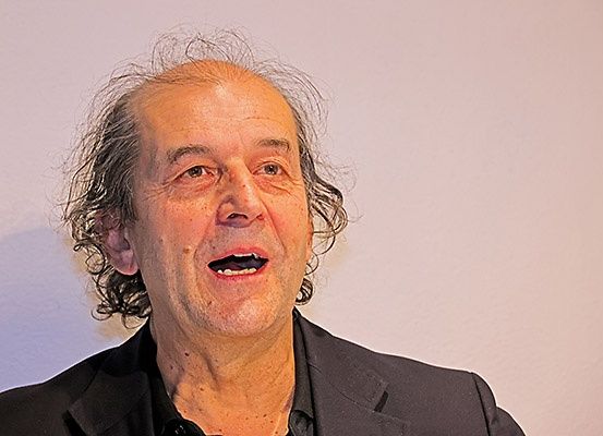 Ein älterer Mann mit lockigem Haar und einem schwarzen Anzug steht vor einem weißen Hintergrund und lächelt.