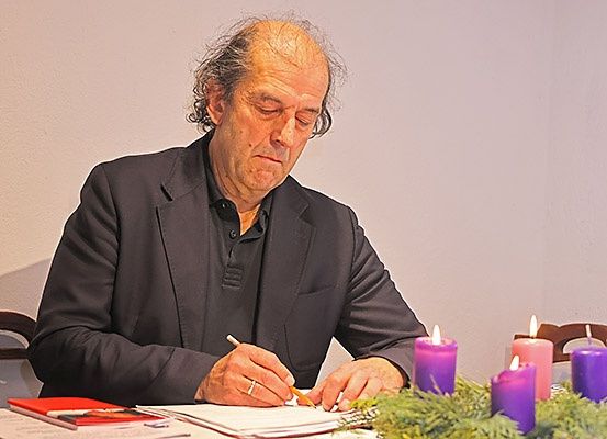 Ein älterer Mann sitzt an einem Tisch und schreibt auf einem Stück Papier. Um ihn herum stehen Kerzen und Kränze.
