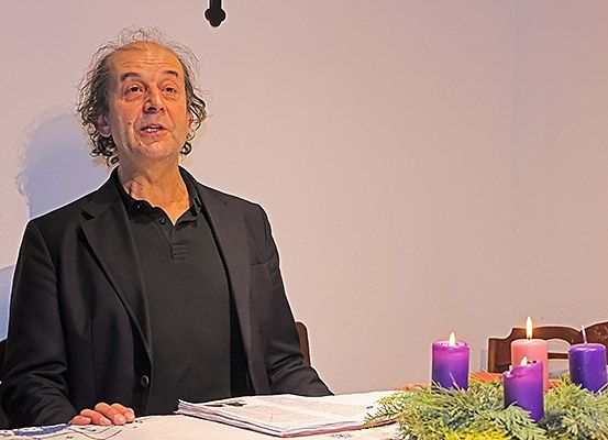 Ein Mann sitzt an einem Tisch mit Papieren und einer Girlande. Er sieht ernst aus. Hinter ihm hat eine weiße Wand drei brennende lila Kerzen.