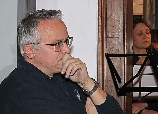 Ein älterer Mann mit Brille sitzt mit der Hand vor dem Mund und schaut auf einen Notenständer mit Noten. Hinter ihm schaut eine Frau ebenfalls nach unten, und ein Mann ist hinter ihr.