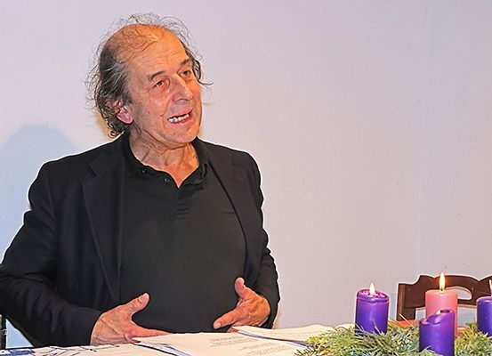 Ein älterer Mann sitzt an einem Tisch mit vier lila Kerzen und Papieren vor ihm. Er trägt ein schwarzes Hemd und eine Jacke und lächelt. Er hat einen Schatten an der Wand hinter ihm.
