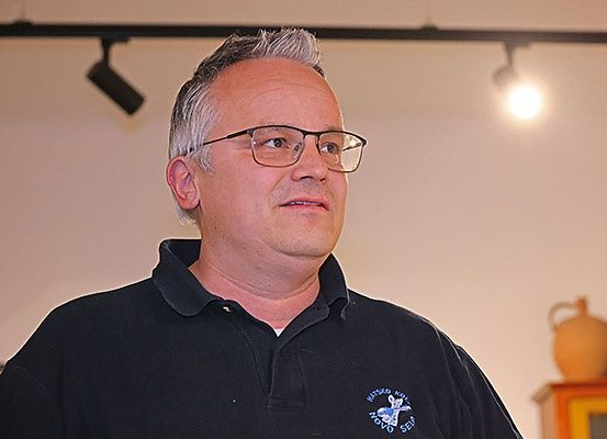 Ein Mann in einem schwarzen Poloshirt mit Brille steht und schaut nach rechts. Hinter ihm befindet sich eine Wand mit einer Einbauleuchte und ein Holzschrank mit einer Vase.