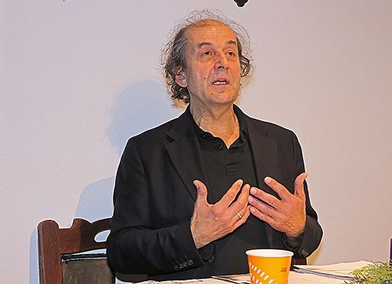 Ein älterer Mann in einem schwarzen Hemd spricht an einem Tisch und gestikuliert mit den Händen. Ein gelber Becher und eine Kerze stehen auf dem Tisch. Der Hintergrund ist eine schlichte Wand.