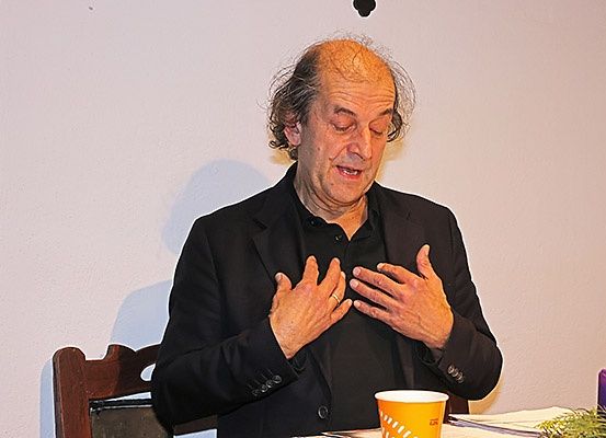 Ein älterer Mann sitzt an einem Tisch, die Hände gefaltet, und schaut nach oben, als ob er betet. Eine lila Kerze ist angezündet auf dem Tisch, und ein gelber Becher steht in der Nähe.