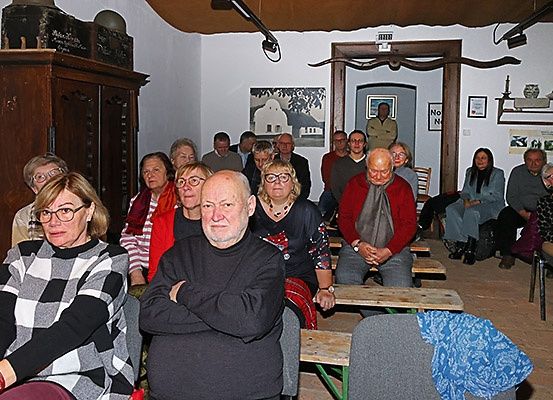 Eine Gruppe von Menschen sitzt in einem Raum mit Holzbänken und Stühlen, einige tragen Brillen und Schals. Eine Person steht in der Tür.