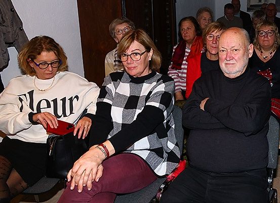 Mehrere Menschen sitzen in einem Raum zusammen, tragen Brillen und sind in verschiedene Winterkleidung gekleidet. Eine Frau vorne trägt einen weißen Pullover mit schwarzen Buchstaben.