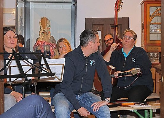 Eine Gruppe Erwachsener sitzt zusammen in einem Raum, wobei zwei Personen Gitarre spielen und eine andere Noten liest. Eine Puppe in einer Vitrine ist im Hintergrund sichtbar.
