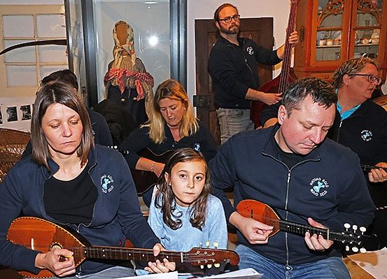 Eine Gruppe von Erwachsenen und Kindern spielt Saiteninstrumente, darunter eine Mandoline und einen Cello, in einem Raum mit Schrank und Fenster.