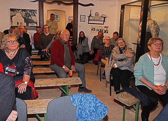 Eine Gruppe von Menschen sitzt auf Bänken und Stühlen in einem Raum. Einige tragen Brillen und Halsketten. An der Wand befindet sich ein Holzkreuz.