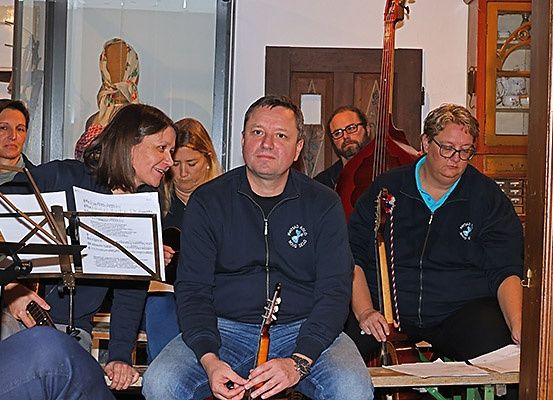 Eine Gruppe von Menschen, darunter ein Mann mit einer Gitarre, sitzt in einem Raum mit Musikinstrumenten. Sie bereiten sich wahrscheinlich auf eine Aufführung vor.