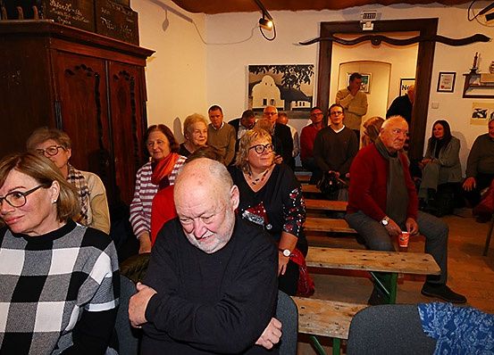 Eine Gruppe von Menschen sitzt auf Holzbänken in einem Raum. Einige tragen Brillen, und ein Mann hält einen roten Becher. Der Raum hat einen Holzschrank, Lampen und gerahmte Bilder an den Wänden.