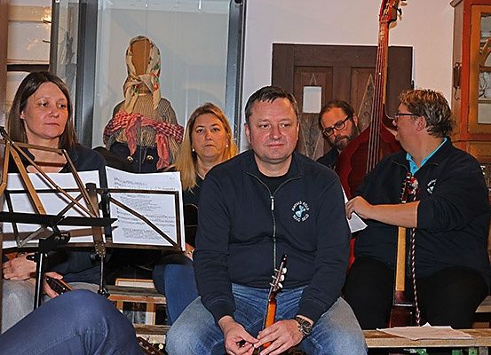 Eine Gruppe von Menschen sitzt mit Instrumenten um sich herum, darunter Gitarren und ein Cello, während eine andere Papiere hält. Sie sind in einem Raum mit einer Holztür und einem Schrank mit Glastüren.