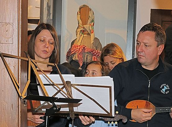 Eine Gruppe von Musikern, darunter eine Frau und ein Mann, spielen Instrumente in einem Raum mit einer Holztür. Die Frau hält einen Notenständer, und der Mann spielt eine Balalaika.