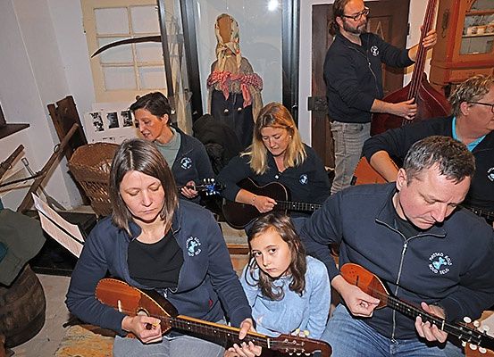 Eine Gruppe von Musikern spielt in einem Raum Instrumente, wahrscheinlich einem Musikraum. Einige von ihnen sitzen auf dem Boden, während andere stehen. Sie tragen alle Jacken. Vor ihnen steht ein Fass, ein Korb und eine Glastür. Dahinter befindet sich ein Schrank mit Glastüren.