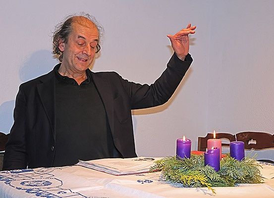 Ein Mann in einem schwarzen Mantel lächelt, während er an einem Tisch mit einem Kranz und vier lila Kerzen sitzt. Hinter ihm sind eine helle Wand und zwei Stühle zu sehen.