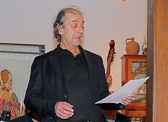 Ein älterer Mann steht drinnen, hält ein Papier in der Hand, mit einem Cello und einer alten Vase auf einem Regal im Hintergrund.