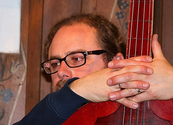 Ein Mann mit Brille und Bart hält eine rote Gitarre in den Händen. Er scheint die Gitarre zu spielen oder zu justieren.