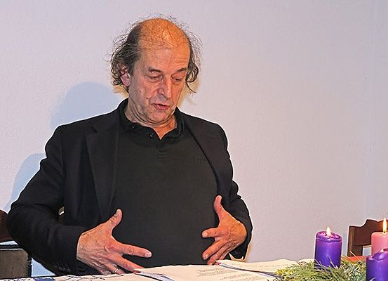 Ein älterer Mann sitzt an einem Tisch mit Kerzen und einem Kranz. Er schaut nach unten, möglicherweise liest oder denkt er.