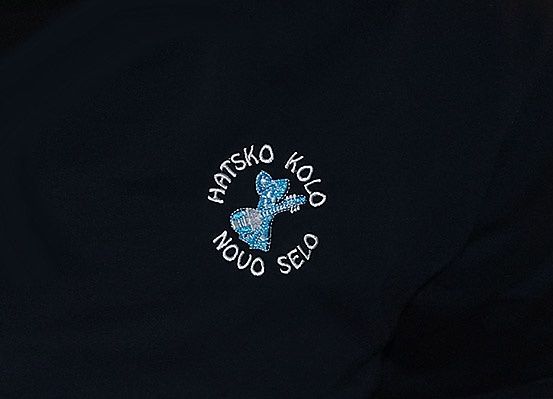 Ein schwarzer Stoff mit einem blauen und weißen Logo, das eine Gitarre zeigt, mit den Worten 'HATSKO KOLO' darüber und 'NOUO SELO' darunter.