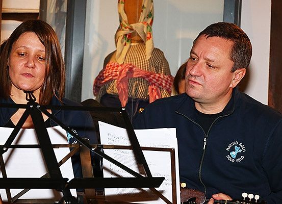Ein Mann und eine Frau bereiten sich auf eine musikalische Aufführung vor. Die Frau hält Notenblätter und der Mann spielt Gitarre. Im Hintergrund ist eine Puppe in traditioneller Kleidung zu sehen.