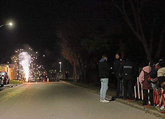 Eine nächtliche Straßenszene mit Feuerwerk. Mehrere Personen, darunter zwei Polizeibeamte, stehen auf dem Bürgersteig. Eine Menge beobachtet von Barrieren geschützt. Bäume und Straßenlaternen sind sichtbar.