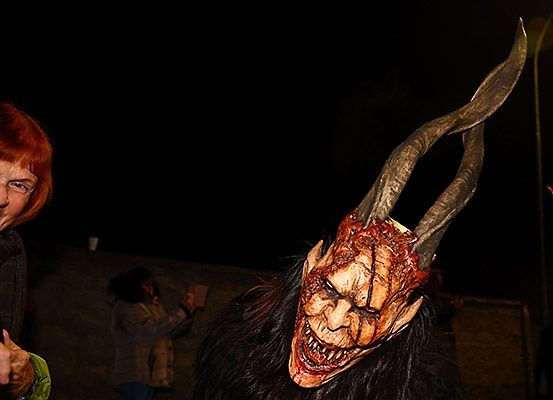 Eine Person mit einer gehörnten Maske steht vor einer Frau mit Brille. Dahinter hält eine weitere Frau eine Kamera in der Hand. Der Hintergrund ist dunkel.