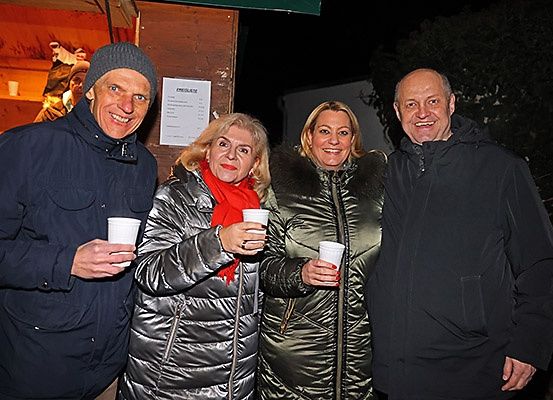 Vier Menschen stehen zusammen in der Dunkelheit und halten Tassen. Der Mann auf der linken Seite trägt eine Mütze und eine Jacke. Die beiden Frauen tragen Jacken mit Pelzbesatz. Der Mann auf der rechten Seite trägt eine schwarze Jacke. Sie lächeln.