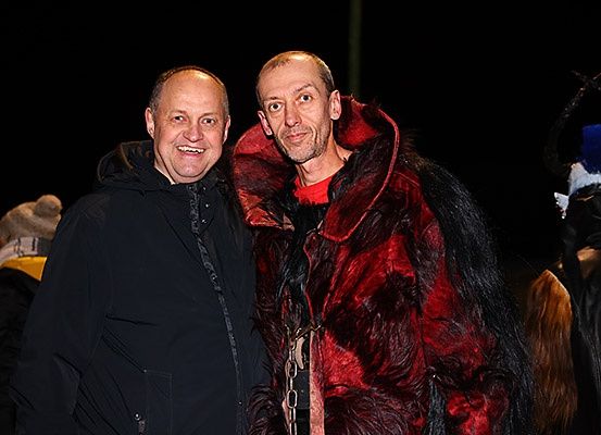 Zwei Männer in Mänteln stehen nah beieinander und lächeln. Einer trägt einen roten Pelzmantel, der andere einen schwarzen Mantel. Hinter ihnen sind andere in Kostümen gekleidet.