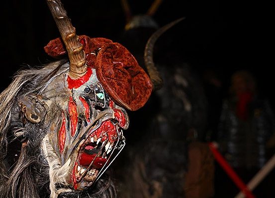 Eine gruselige Maske mit Hörnern und roter Farbe, die von einer Person in einem Kostüm getragen wird. Sie wird von einem dunklen Hintergrund beleuchtet.