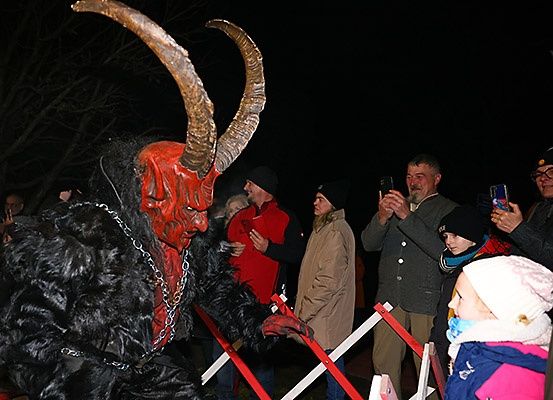 Ein Mann in Teufelskostüm mit Hörnern begrüßt Menschen bei Nacht. Sie machen Fotos mit ihren Handys.