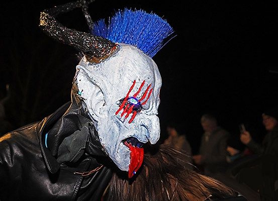 Eine Person in einer dämonischen Maske mit blauen Haaren und roten Augen steht im Dunkeln. Im Hintergrund sind mehrere Personen zu sehen.