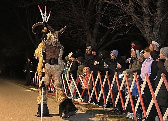 Eine Person in einem Ziegenkostüm steht bei Nacht auf der Straße, umgeben von Zuschauern. Einige Leute machen Fotos.