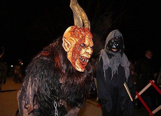 Zwei Personen in Kostümen gehen im Dunkeln. Einer trägt eine Hörnermaske und ein schwarzes Outfit, der andere eine graue Kapuze. Rechts steht eine rote Barriere.