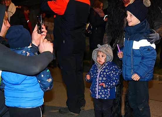 Ein Mann macht ein Foto mit einem Handy von zwei Kindern, eines trägt einen pelzigen Hut, und einem Mann in einem Kostüm, alles in einer verschneiten Außenumgebung.