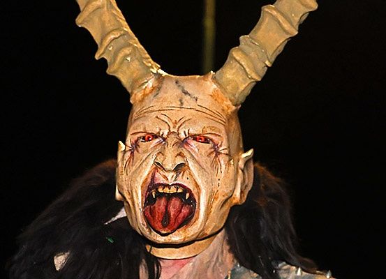 Eine Person trägt eine Maske mit Hörnern und einem grotesken Gesicht. Die Maske hat einen schwarzen Hintergrund und rote Augen.
