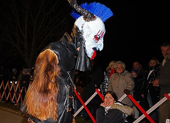 Eine Person in einem Kostüm mit gehörntem Maske und Fell steht bei Nacht vor einer Menge. Dahinter schauen Leute zu, einige tragen Brillen. Ein roter Barriere trennt sie.