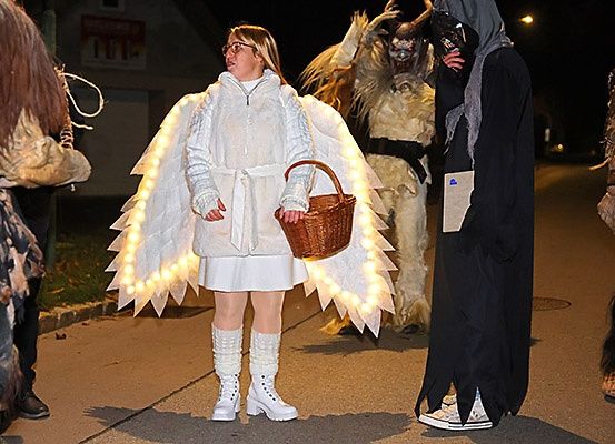 Eine Frau in Engelsflügeln und mit einem Korb geht auf der Straße mit einer Gruppe von Menschen in Kostümen.