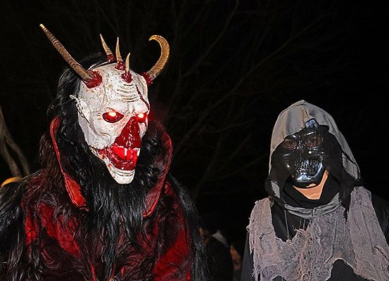 Zwei Personen in dunklen Kostümen, eine trägt eine rote Dämonenmaske mit Hörnern und leuchtenden Augen, die andere ein schwarzes Kapuzenkostüm mit Maske, stehen in einer schwach beleuchteten Outdoor-Umgebung.