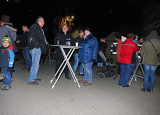 Eine Gruppe von Menschen steht nachts draußen. Einige halten Tassen. Ein Mann lächelt. Sie stehen vor einem Tisch mit Flaschen. Ein Kinderwagen ist in der Nähe.