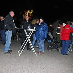 Eine Gruppe von Menschen steht nachts draußen. Einige halten Tassen. Ein Mann lächelt. Sie stehen vor einem Tisch mit Flaschen. Ein Kinderwagen ist in der Nähe.