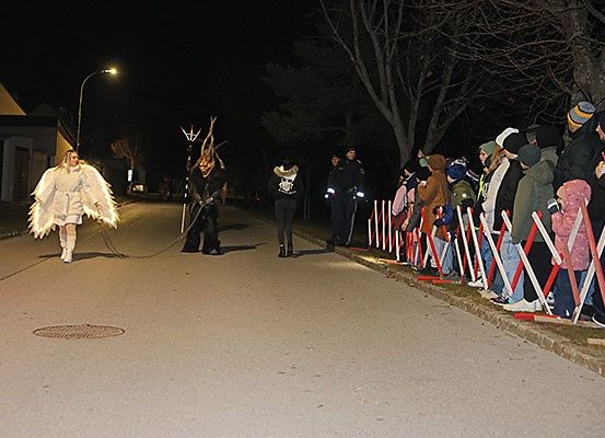 Eine Nachtveranstaltung mit einer Person, die als Engel verkleidet ist und die Straße entlanggeht, einer Person mit Maske und Leuten, die hinter einer Barriere zuschauen.
